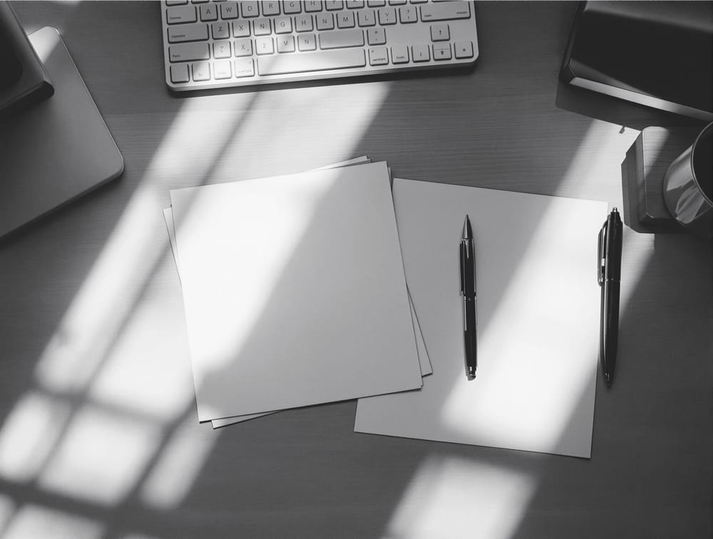 An uncluttered desk photographed from above with loose blank sheets and a pen in morning light.